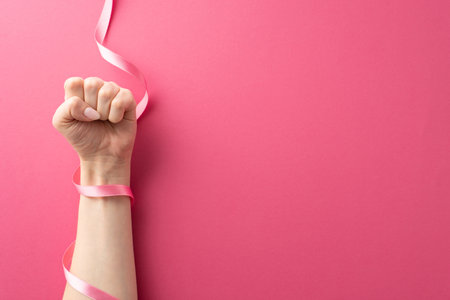 Spreading Breast Cancer Awareness Month message. Overhead shot of female hand in a fist and pink ribbon on a magenta pink isolated setting, suitable for text or promotional materialsの写真素材