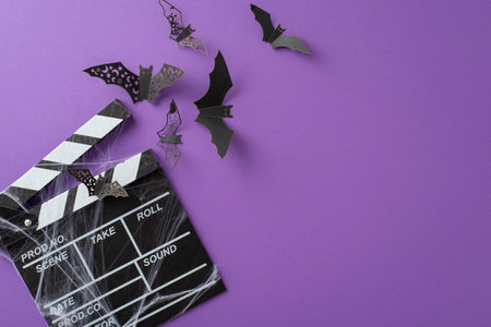 Prepare for an unforgettable Halloween night with a captivating visual of film clapper and Halloween-themed paper bats against purple isolated background, copyspace ideal for your marketing needs