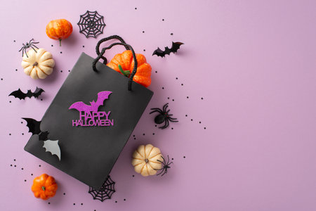 Share Halloween joy with thoughtful gifts. Seen from above, a black paper gift bag adorned with Halloween decor rests on pastel purple isolated background, offering copy-space for ads or textの写真素材