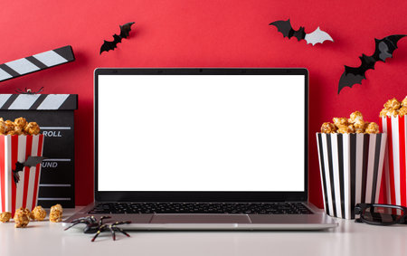 Create your Halloween cinema at home with side view of laptop, eerie decor, spiders, bats, 3D glasses, clapperboard and popcorn boxes against a red wall with blank space for promotion or advertisingの写真素材