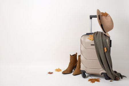 Female autumn adventure with luggage. Side view of a chic suitcase, comfy scarf, ankle boots, felt hat, pine cone, and maple leaves against a white background with space for text or adsの写真素材