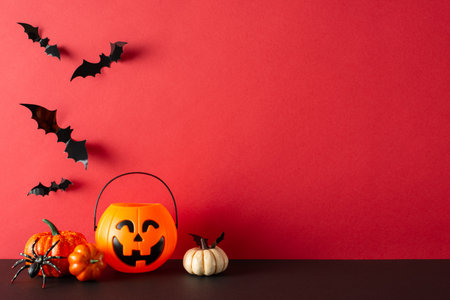 Spooky Halloween-themed decor for festive vibe. Side view of table adorned with pumpkins, jack-o'-lantern basket, spider, eerie bat silhouettes against red wall backdrop, leaving room for text or adsの写真素材