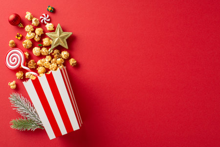 Deck the halls for New Year's film night: striped cardboard bucket with delicious popcorn, baubles, star decor, festive lollipop, candies, frosted fir branch, and a red backdrop with text spaceの写真素材