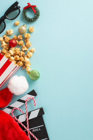 Christmas-themed filmstrip with holiday treats concept. Top view vertical photo of festive movie clapper, 3D glasses, sweet candy canes, Santa's hat, ornaments, tiny wreath, popcorn on snowy backdropの写真素材