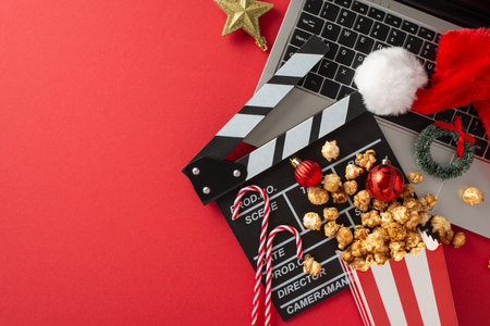 Create a festive atmosphere for your at-home movie night. Top view of a laptop, popcorn, Santa's hat, baubles, star, tiny wreath, candies, clapper, empty space for your message against red backgroundの写真素材