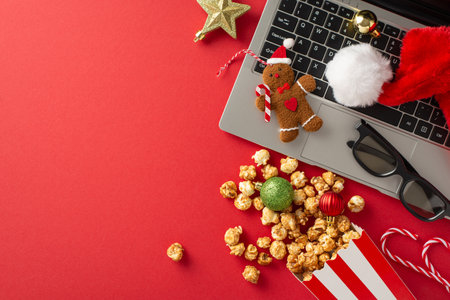 Get into the New Year mood with cozy movie night at home. Top view of a laptop, 3D glasses, tasty popcorn, Santa's hat, ornaments, star decor, gingerbread man toy and a red backdrop for your textの写真素材