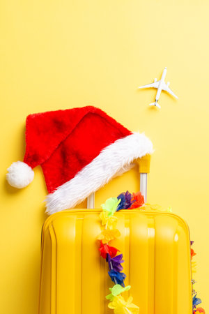 Island-bound for New Year's! Top-view vertical photo featuring suitcase, Santa hat, floral garland, and a miniature plane on a vibrant yellow backdrop. Get ready for a tropical celebrationの写真素材