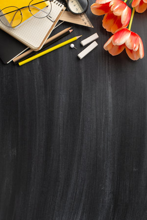 Mark sophisticated Teacher's Day concept. Vertical top view capturing classroom necessities, notebooks, pencils, chalk, beautiful tulip bouquet on blackboard setting, area for messages or promotionsの写真素材