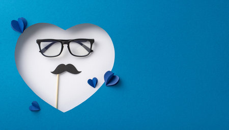 Creative Father's Day concept featuring a white heart with glasses and a mustache prop on a blue background, symbolizing parental loveの写真素材
