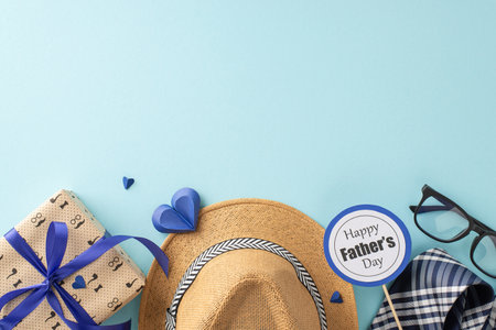 Personalize your Father's Day message with a top-view arrangement highlighting a straw hat, elegant necktie, gift box, glasses, postcard, and paper hearts on a soothing pastel blue sceneの写真素材