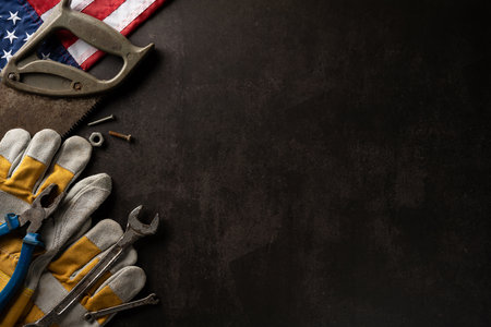 Flat lay of assorted tools with protective gloves and American flag on dark background, symbolizing...