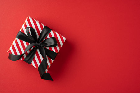 A beautifully wrapped gift box with red and white stripes and a black ribbon set against a red backdrop. Perfect for celebrating holidays and special occasionsの写真素材