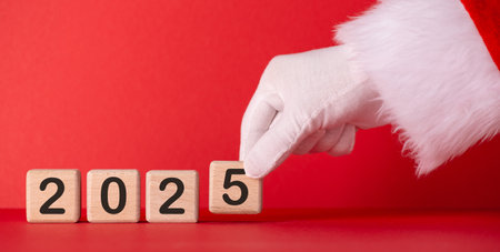 A gloved hand in a Santa suit flips wooden blocks displaying the year 2025 against a bright red background, symbolizing the New Year countdown and holiday excitementの写真素材