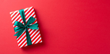 A beautifully wrapped Christmas gift with a green ribbon and striped paper on a vibrant red background, symbolizing festive joy and holiday spiritの写真素材