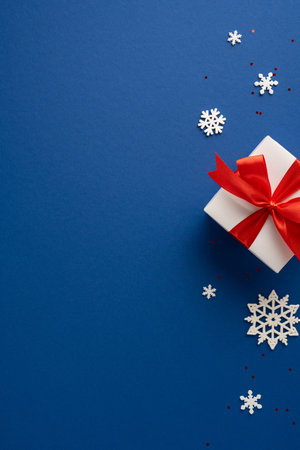 White vertical gift box with a striking red ribbon surrounded by decorative snowflakes on a blue background, creating a festive and elegant Christmas atmosphereの写真素材