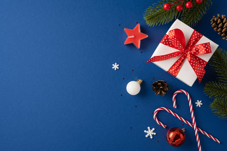 A beautifully wrapped Christmas gift with a red ribbon surrounded by holiday decorations on a blue background. Includes candy canes, ornaments, and pine branchesの写真素材