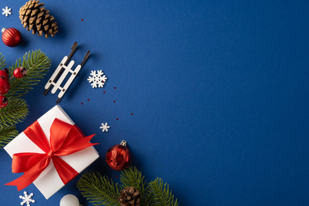 Decorative Christmas scene featuring a white gift box with red ribbon, surrounded by festive ornaments, pine branches, and a sled on a navy blue backgroundの写真素材