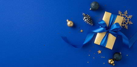 Gold gift box with blue ribbon surrounded by festive ornaments on a blue backdrop, conveying elegance and holiday spiritの写真素材