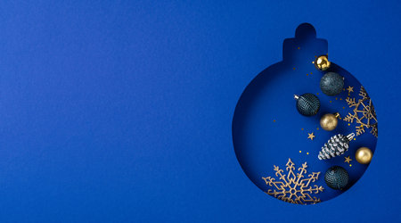A creative Christmas design featuring an ornament-shaped cutout of Christmas ball filled with festive decorations and golden snowflakes on a dark blue backgroundの写真素材