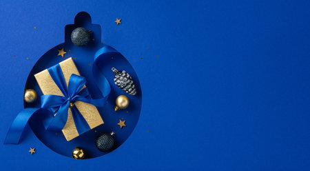 A beautiful top view of Christmas present wrapped in gold paper with a blue ribbon, surrounded by festive ornaments and stars on a blue background. Perfect for holiday celebrations and gift-giving ideasの写真素材