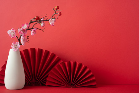 A vibrant red backdrop featuring a white vase with cherry blossom branches, complemented by red paper fans. Perfect for celebrating the Chinese New Year 2025 with cultural eleganceの写真素材