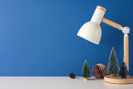 Minimalist side view Scandinavian desk scene featuring a modern lamp and decorative mini Christmas trees against a blue wall, capturing cozy winter vibesの写真素材