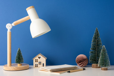A side view of minimalist winter-themed desk with a Scandinavian design, featuring a lamp, notebook, wooden house, and mini Christmas trees against a blue backgroundの写真素材