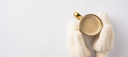 Hands wearing warm mittens hold a gold-rimmed coffee cup, creating a cozy winter atmosphere. The minimal white background emphasizes the warmth and comfort of the scene.の写真素材
