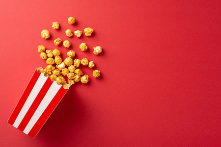 Popcorn spills from a classic red and white striped box on a vibrant red background, creating a dynamic and eye-catching visual perfect for movie night themesの写真素材