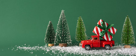 A small red toy truck carrying a wrapped Christmas gift next to miniature snowy trees on a green background. Ideal for holiday and festive conceptsの写真素材