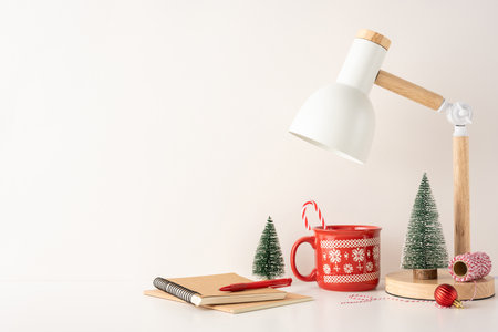A side view of minimal Scandinavian workspace featuring a desk lamp, festive red mug, tiny Christmas trees, and stationery. The scene evokes warmth and holiday spirit in a simple and elegant styleの写真素材