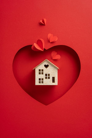 A vertical small house model with a heart window is displayed on a heart shaped red background, surrounded by paper hearts. Perfect for themes of love, home, and Valentine's Dayの写真素材