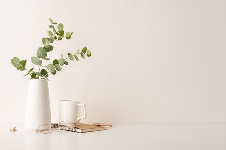 A minimal desk setup featuring a white vase with eucalyptus, a notebook, glasses, and a mug. Perfect for concepts of simplicity, organization, and a serene workspace environmentの写真素材