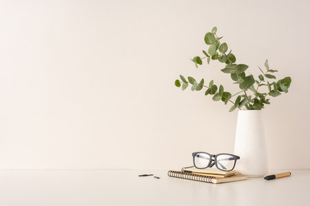 Minimalist desk setup with glasses, a notebook, pen, and a vase of eucalyptus leaves in a peaceful, natural design. Ideal for themes of productivity, elegance, simplicity, and contemporary workspace aestheticsの写真素材