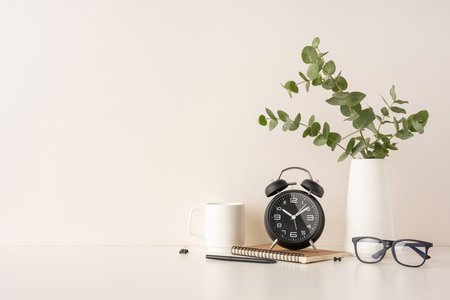 Minimalist desk arrangement featuring a black alarm clock, glasses, notebook, and vase holding eucalyptus. Perfect for modern office inspirations or productivity themesの写真素材