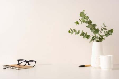 A composition showing a serene workspace setup with a notebook, glasses, eucalyptus, and white ceramic items on a white surface, providing a minimalist and tidy aestheticの写真素材
