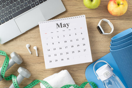 A calendar alongside fitness accessories like weights, smartwatch, yoga mat, and measuring tape on a wooden deskの写真素材