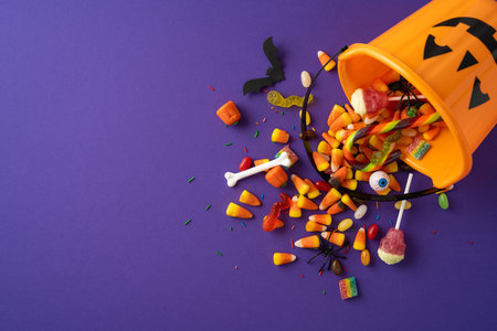 Orange pumpkin bucket with various colorful candies spread out on a purple surface
