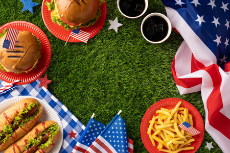 Festive picnic setup celebrating Labor Day and Independence Day with burgers, hot dogs, fries, and American flag decorations on a grassy settingの写真素材