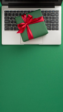 A small vertical green gift box with a red ribbon sits on a laptop keyboard, surrounded by a solid green background. Perfect for holiday, gift, or celebration conceptsの写真素材