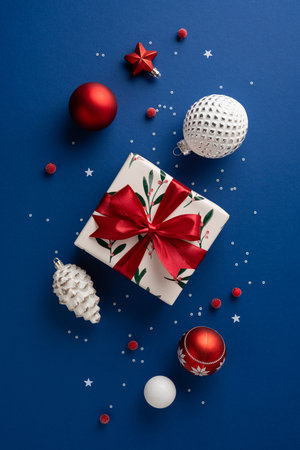 A vertical holiday scene featuring a wrapped gift, red and white ornaments, pinecone, and confetti on a blue backdrop. Perfect for Christmas, New Year greetings, gift ideas, and festive promotionsの写真素材