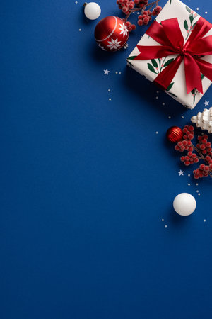 A vertical festive scene featuring a wrapped gift with a red ribbon, red ornament, white balls, and berry sprigs on a blue backdrop. Perfect for Christmas, New Year, greeting cardsの写真素材