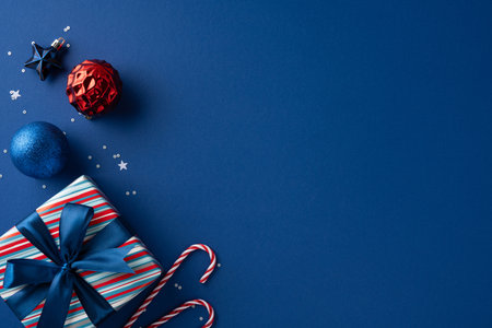 A festive scene featuring a striped gift box with blue ribbon, blue and red ornaments, and candy canes scattered on a dark blue backdrop to evoke holiday cheerの写真素材