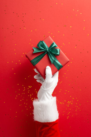 A vertical white-gloved hand holds a red gift box tied with a green satin ribbon against a red backdrop with gold confetti, conveying joy, generosity, and holiday celebrationの写真素材