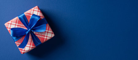 A wrapped gift box with red plaid pattern and a bright blue ribbon rests on a dark blue surface, evoking festive holiday spirit, gift giving, and seasonal greetings for Christmas and New Yearの写真素材