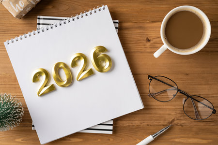 A desk scene with a white notebook displaying gold 2026 digits, a cup of coffee, round glasses, and a pen, capturing festive preparation and anticipation for New Year greetingsの写真素材