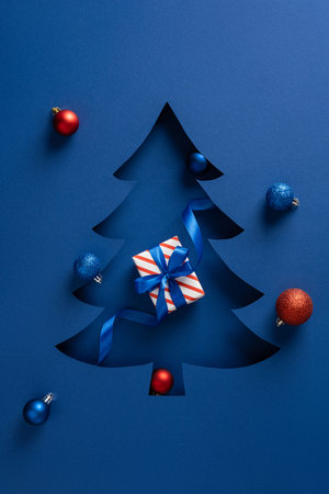 Blue vertical Christmas tree cutout on a dark blue background features a striped gift, blue ribbon, and colorful ornaments, capturing holiday spirit for Christmas and New Year campaignsの写真素材
