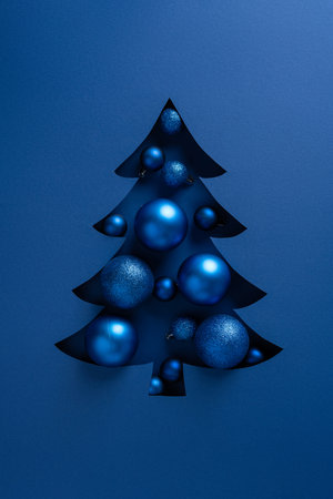 Blue vertical Christmas tree cutout reveals shiny ornaments in shades of blue against a simple blue background, evoking festive holiday spirit and a New Year greetingの写真素材