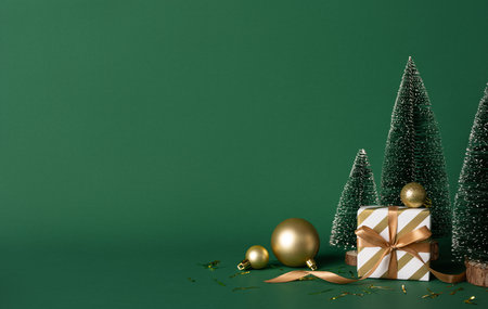 A stylish Christmas scene featuring a gold ribbon wrapped gift, tiny frosted trees, and gold ornaments set against a rich green backdrop, conveying festive warmth and New Year anticipationの写真素材