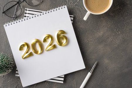 A modern desk scene with a white notebook featuring gold 2026 digits, a cup of coffee, glasses, and...